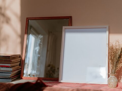 Two thin wooden picture frames leaning against a peach wall in a lived-in interior, featuring a blank mockup template alongside a stack of books and dried flora for art or display