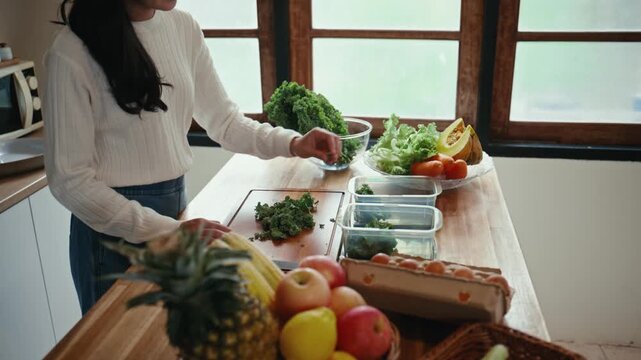 Young asian woman chopping kale on a wooden cutting board in her kitchen. She is preparing fresh ingredients for a healthy meal at home