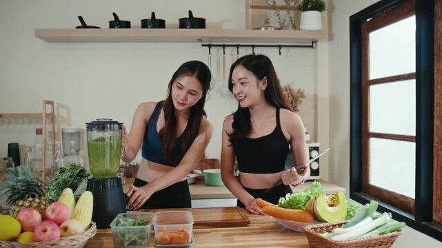 Two smiling Asian women in sportswear preparing a healthy drink. One is using a blender while the other holds a tablet, checking the recipe