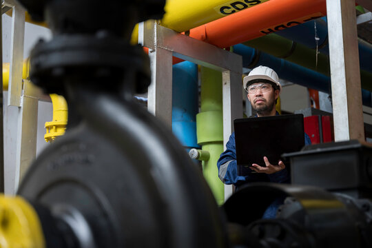 Focused professional male industrial engineer uses a laptop computer to inspect heavy machinery and colorful utility pipes during maintenance inside a modern manufacturing plant facility.