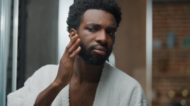 Portrait African American man touching gentle face skin beard grooming looking at mirror reflection wearing bathrobe at bathroom handsome ethnic guy smiling bearded male touch facial hairstyle smile