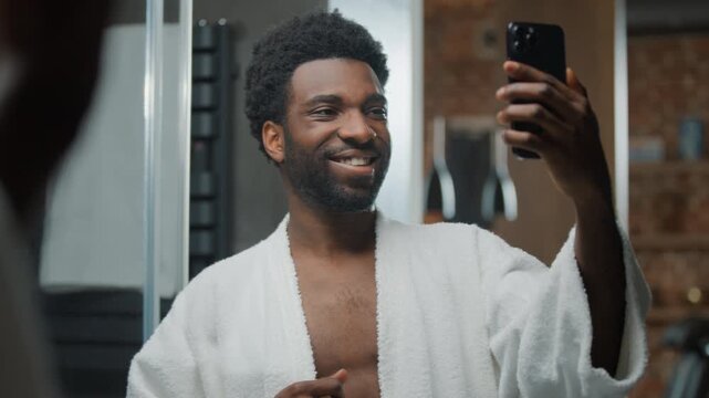 Happy African American man influencer smiling posing for selfie photo at bathroom mirror reflection ethnic guy male smile for mobile phone picture video smartphone chat vlog shooting skin care routine