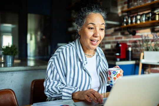Older woman using laptop and drinking coffee in home kitchen