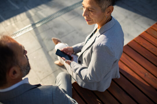 Senior businesswoman with smartphone talking to colleague on outdoor bench