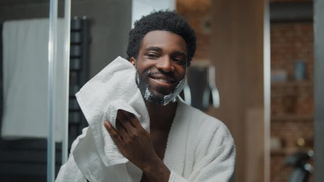 Mirror reflection of happy positive smiling African American man wiping cheeks after shaving shave gel foam soap optimistic male guy wipe foamy beard with white towel finishing trimming in bathroom
