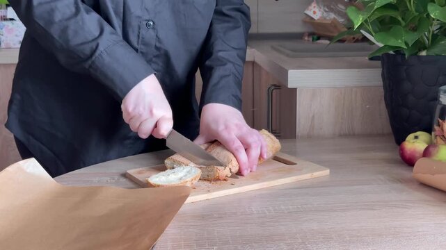 Slicing fresh loaf of bread on wooden cutting board with kitchen knife during homemade food preparation in home kitchen, close-up cooking process with human hands