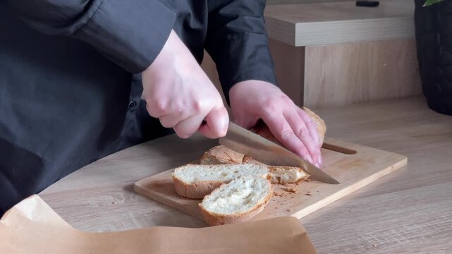 Slicing fresh loaf of bread on wooden cutting board with kitchen knife during homemade food preparation in home kitchen, close-up cooking process with human hands