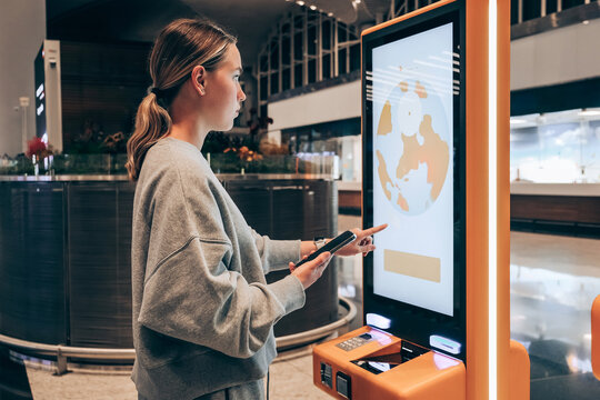 woman using a self-service check-in kiosk and smartphone at a modern airport, illustrating contactless travel, digital passenger services and seamless airport technology