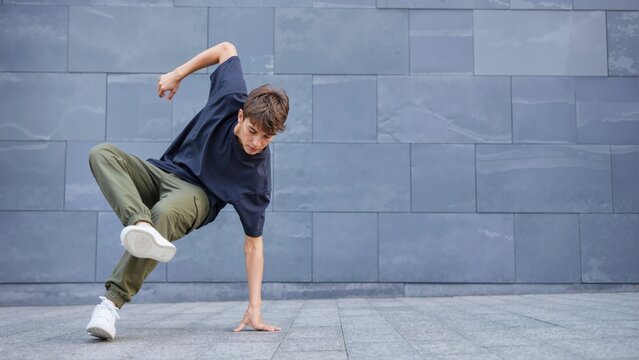 Teen boy performing a street dance move against a grey tiled wall. Young breakdancer balancing on one hand in an urban setting. Active lifestyle and youth hobby concept