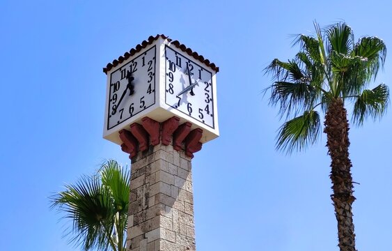 Low-angle view of the historic stone Clock Tower at Kanari square in Zea Marina, Piraeus, Greece, framed by palm trees against a clear blue sky. 
