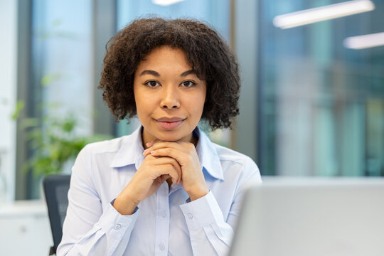 Close-up portrait of a young African American woman sitting in an office near a laptop, folding her hands together and looking confidently at the camera
