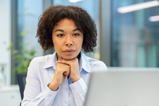 Close-up photo of a young African American businesswoman sitting in an office at her workplace, folding her hands together and looking seriously at the laptop screen