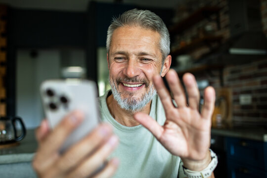 Smiling senior man waving on smartphone video call at home kitchen