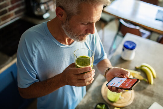 Senior man checking fitness app while drinking green smoothie in home kitchen