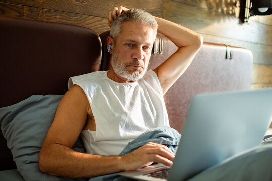 Middle-aged man working on laptop with earbuds in bed at home