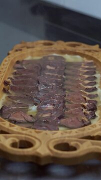 Chef in black gloves pouring hot meat broth from a saucepan over traditional uzbek beshbarmak dish with sliced beef and kazi on a carved wooden platter, vertical video.
