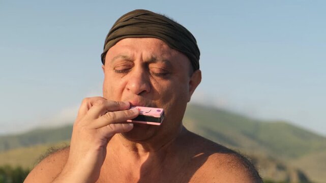 Portrait of a tanned overweight man playing a harmonica shirtless against mountain scenery at sunset. Sad emotional melody. Non European man wearing a headscarf.