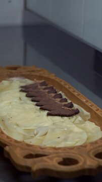 Chef in black gloves placing sliced boiled beef and kazi horsemeat sausage over dough sheets for traditional uzbek naryn beshbarmak dish on a carved wooden plate, vertical video.