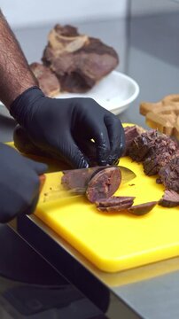 Chef in black gloves slicing traditional uzbek kazi horsemeat sausage and boiled beef on a yellow cutting board, slow motion vertical video.