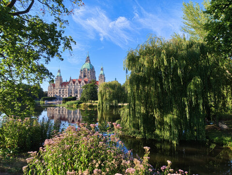View of the New Town Hall reflected in the lake at Maschpark with lush weeping willows and blooming flowers in the foreground in Hannover, Niedersachsen, Germany.