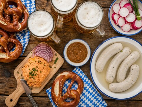 Bayerische Brotzeit aus der Vogelperspektive mit Wei&szlig;w&uuml;rsten, Brezeln, Obatzda, s&uuml;&szlig;em Senf, Radieschen und sch&auml;umenden Bierkr&uuml;gen auf rustikalem Holz.