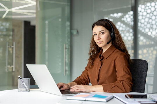 Young female call center agent wearing a headset and brown shirt, working on a laptop at a modern office desk, interacting with customers