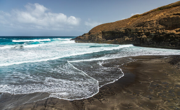 View of the black sand beach at Santo Antao with crashing waves and rugged volcanic cliffs under a partly cloudy sky in Cruzinha, Ribeira Grande, Cabo Verde.