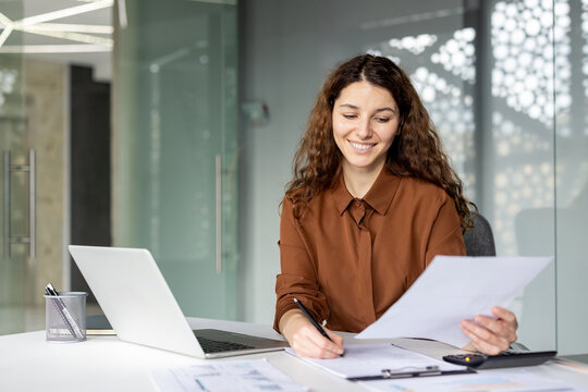 Young businesswoman smiling. Organizing and reviewing financial paperwork while working at a modern office desk with a laptop. Calculator. And various business charts