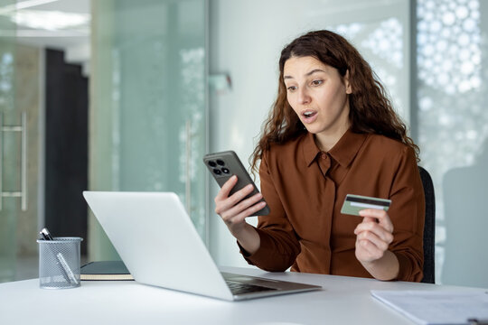 Woman reacting with surprise while holding a smartphone and credit card, using a laptop for online shopping, indicating an unexpected expense or offer during a transaction