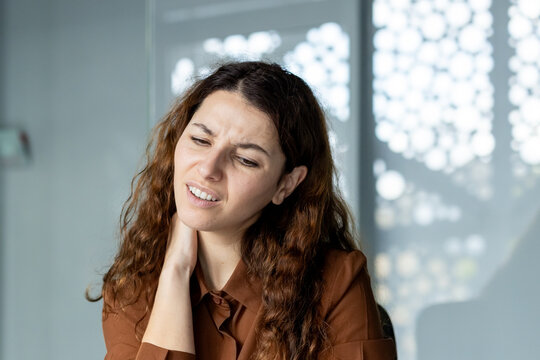 Young woman feeling intense neck pain and muscle tension, suffering from discomfort and headache while working in a modern office environment