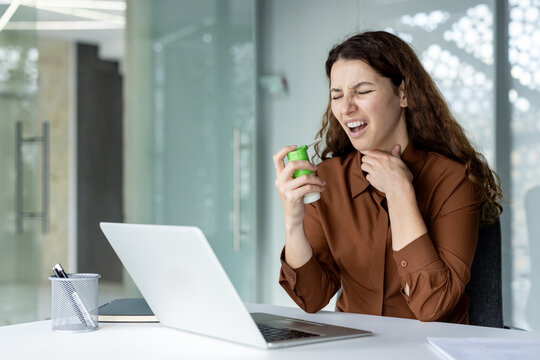 Woman feeling sore throat and pain, using medical spray or inhaler for relief while working at a modern office desk with a laptop