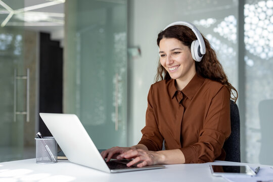 Young businesswoman smiling, wearing white headphones, and typing on a laptop computer while sitting at a desk in a modern office, engaged in remote work or online communication