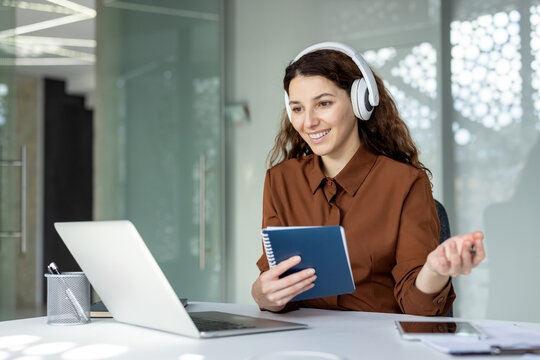 Smiling woman wearing headphones and holding a notebook, participating in a virtual meeting or online education session from an office workspace