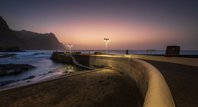 View of the coastal pier at Ponta do Sol on Santo Antao island during sunset with glowing streetlights and waves crashing against the shore in Ponta do Sol, Ribeira Grande, Cabo Verde.