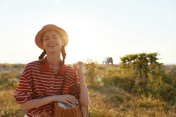 Obraz premium A joyful woman in a striped shirt and straw hat stands in a sunlit field with a woven basket, embracing carefree outdoor lifestyle and warm summer vibes. The scene radiates ease. Warm.