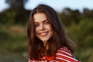 Obraz premium A woman lifestyle portrait outdoors with a bright smile in natural light, relaxed pose, casual striped shirt. A moment of confidence, comfort, and happiness in a sunny meadow.
