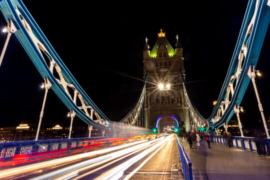 View of Tower Bridge at night with vibrant light trails from passing traffic and pedestrians walking on the sidewalk in London, England, United Kingdom.