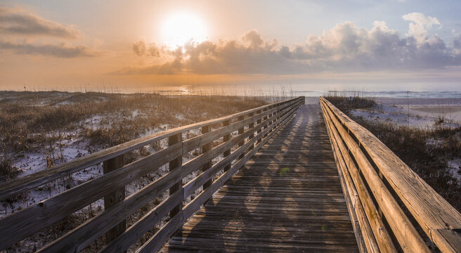 View of a wooden boardwalk leading to the sandy beach at sunrise with sea oats growing in the dunes and calm ocean waves under a warm golden sky in Gulf Shores, Alabama, United States.