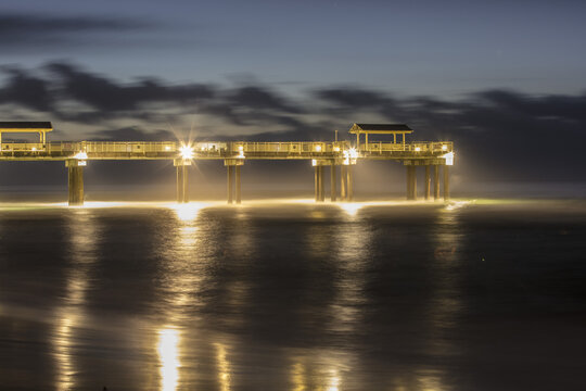 View of the illuminated pier stretching over the calm ocean waters at night with soft reflections on the surface in Gulf Shores, Alabama, United States.