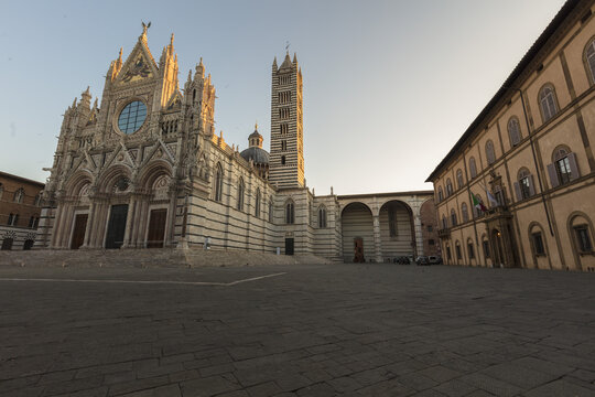 View of the Siena Cathedral with its intricate Gothic facade and bell tower overlooking the paved square during golden hour in Siena, Tuscany, Italy.