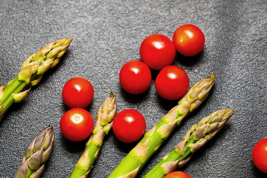Fresh vegetables lay on a dark surface with bright red tomatoes and green asparagus arranged together