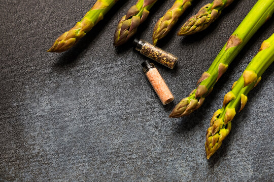 Fresh asparagus spears arranged with small spice containers on a dark surface
