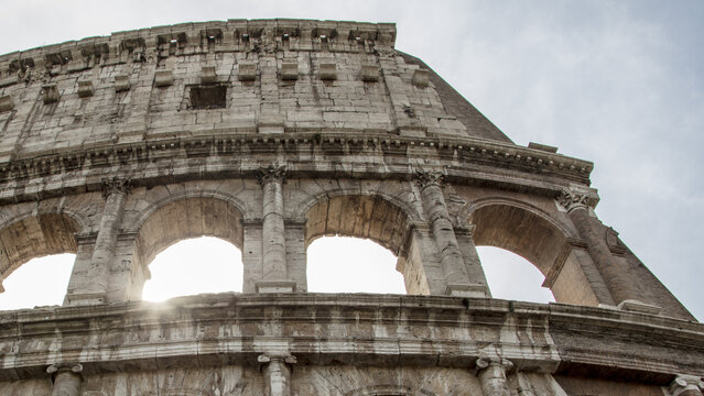 View of the ancient stone arches of the Colosseum in Rome, with sunlight filtering through the historic masonry against a cloudy sky in Roma, Lazio, Italy.