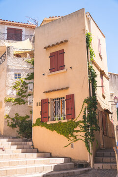 A charming multi-story terracotta building features classic reddish-brown wooden shutters and vibrant green vines climbing its exterior facade. Set along a stepped stone alleyway, this picturesque str