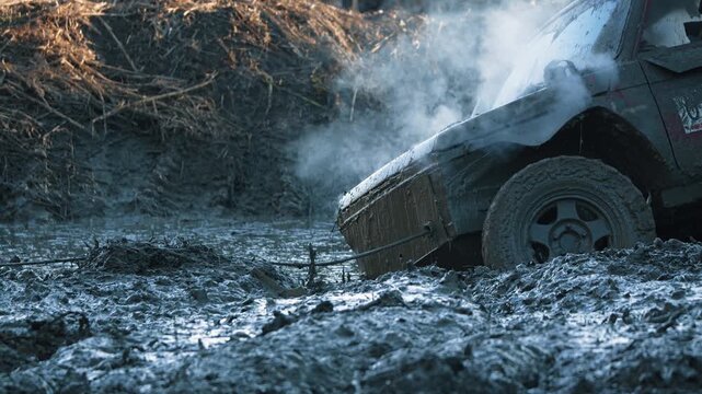 Close up of 4x4 vehicle stuck in thick mud during off road competition. Smoke rises as winch cable pulls car from difficult terrain. Low angle view emphasizes struggle and extreme adventure