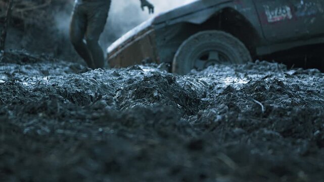 Low angle shot captures textured mud in sharp focus. Out of focus background shows person and 4x4 vehicle stuck. Gritty, cinematic scene with cool blue tones suggests struggle and adventure