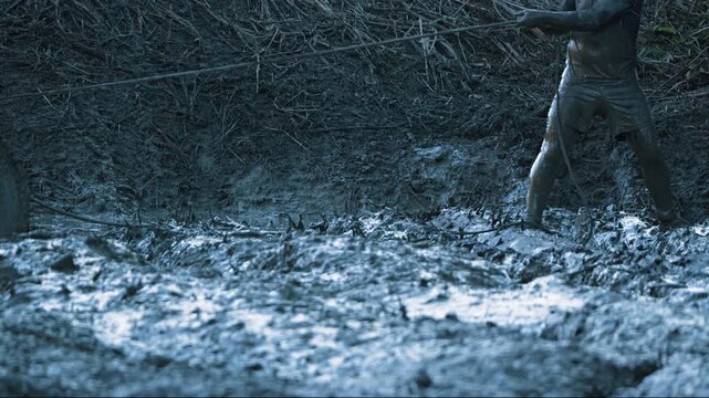 A mud-covered co-pilot stands in deep mud, pulling on a winch cable to free a stuck vehicle. Low-angle shot captures the determination of driver during an extreme off-road adventure. Cold, harsh tones