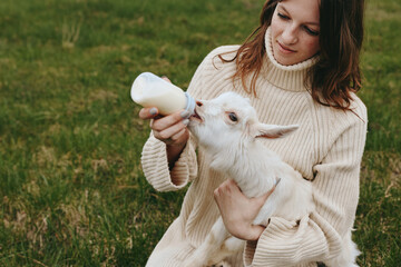 Obraz premium A woman in a cozy sweater bottle feeds a white goat kid outdoors in a rural field, capturing a tender moment of care, bonding, and everyday life with farm animals.