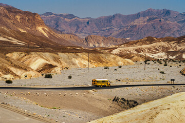 Yellow school bus driving on empty desert highway surrounded by massive eroded mountains, american road trip and education concept © Jakub