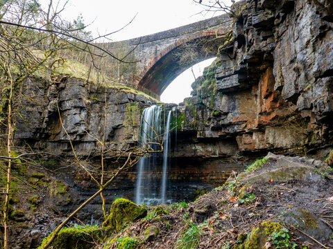 View of Ashgill Force waterfall flowing over dark rocky cliffs beneath an old stone arch bridge, surrounded by mossy rocks and bare trees in Alston, England, United Kingdom.